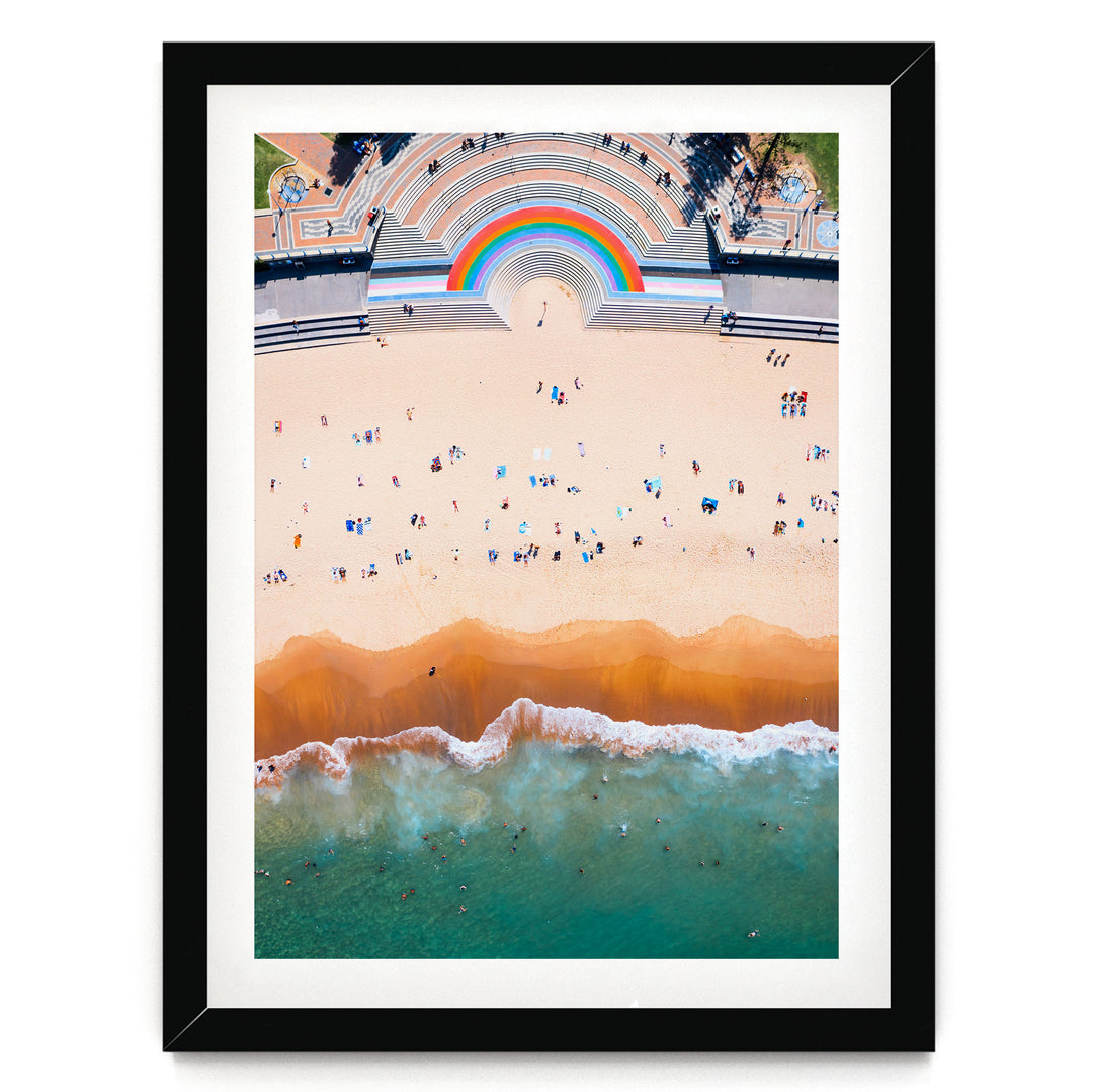 The image is an aerial view of a beach with a large, colorful arch structure in the background, and the ocean with waves crashing onto the shore.