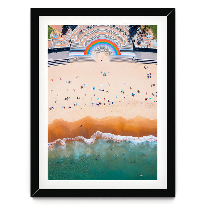 The image is an aerial view of a beach with a large, colorful arch structure in the background, and the ocean with waves crashing onto the shore.
