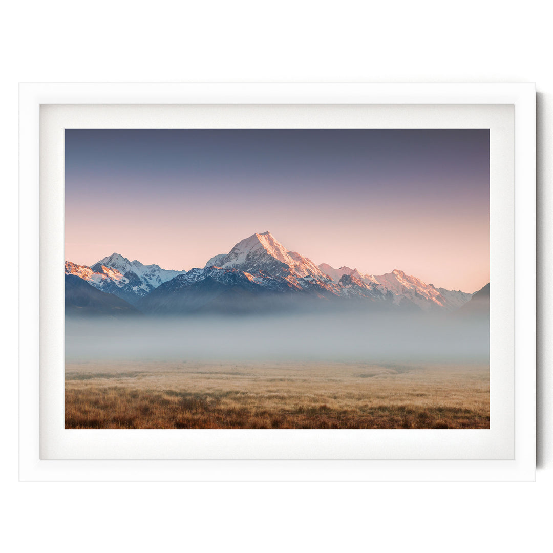 A breathtaking landscape featuring snow-capped mountains in the distance, with a hazy, golden-hued sky above. The scene is framed within a white border, creating a sense of depth and perspective.
