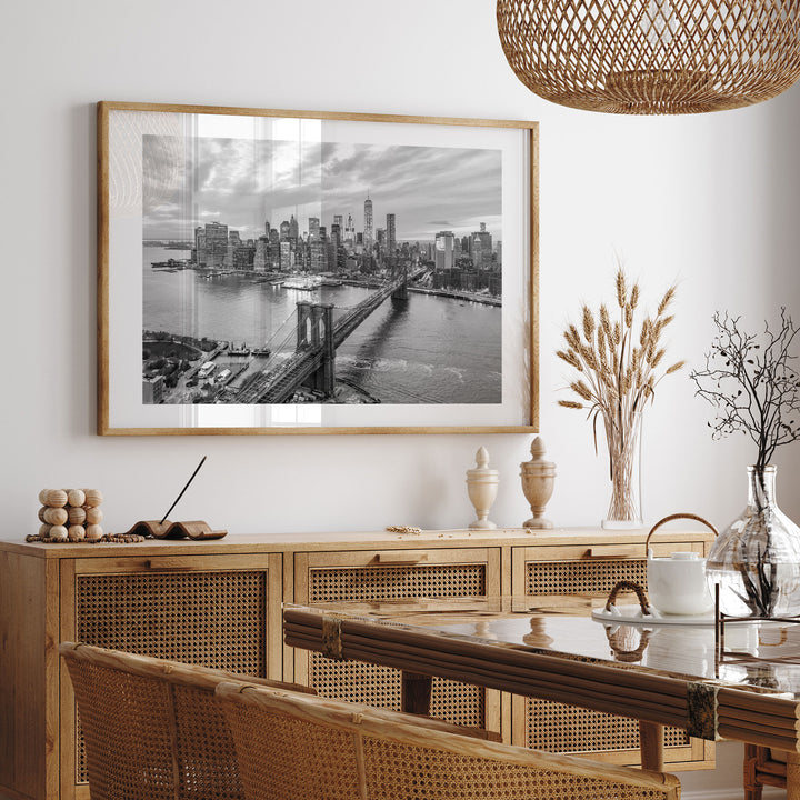 a large framed black and white photograph of the Brooklyn Bridge hanging on a wall above a wooden cabinet. The photograph captures the iconic bridge spanning the East River, with the city skyline visible in the background. The room has a modern and minimalist design, with a woven basket hanging from the ceiling and a vase of dried flowers on a table.