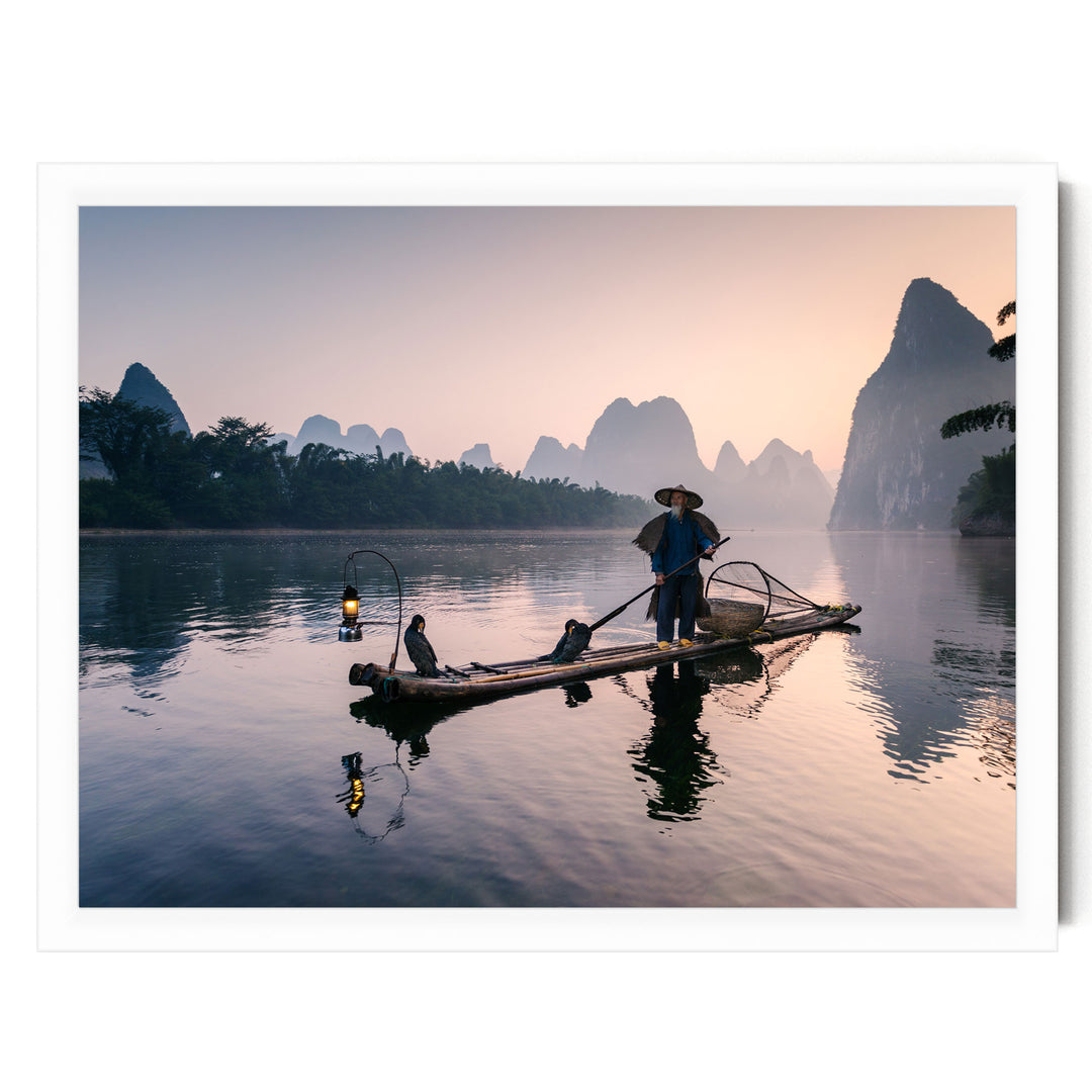 A person in a blue outfit is standing on a long wooden boat, paddling across a calm river at sunset.