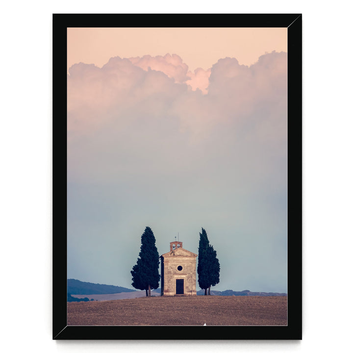 A small stone church with a bell tower sits in a field, surrounded by trees, with a cloudy sky in the background.