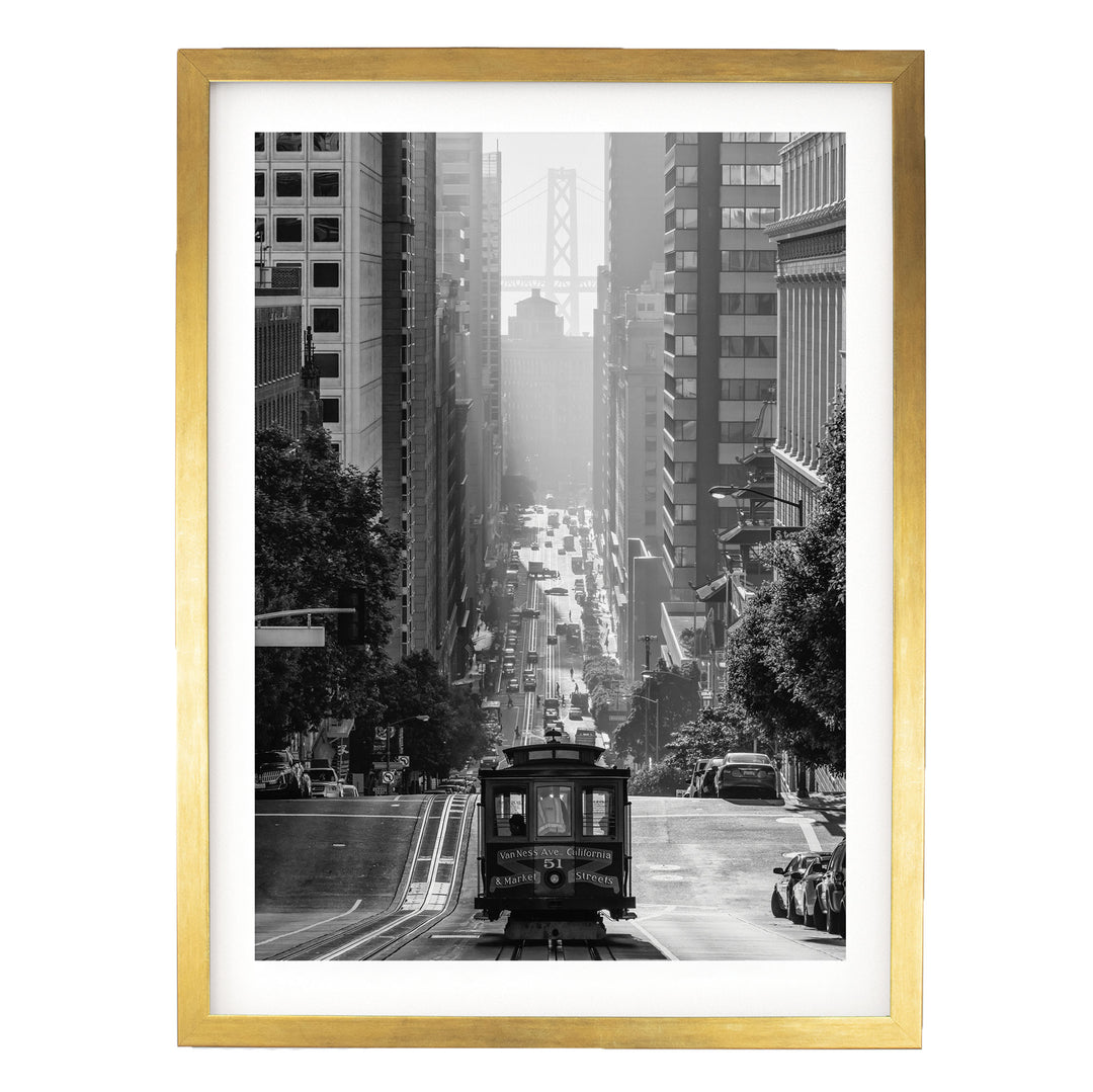 A black and white photograph of a trolley car traveling down a city street, surrounded by tall buildings and a bridge in the distance.