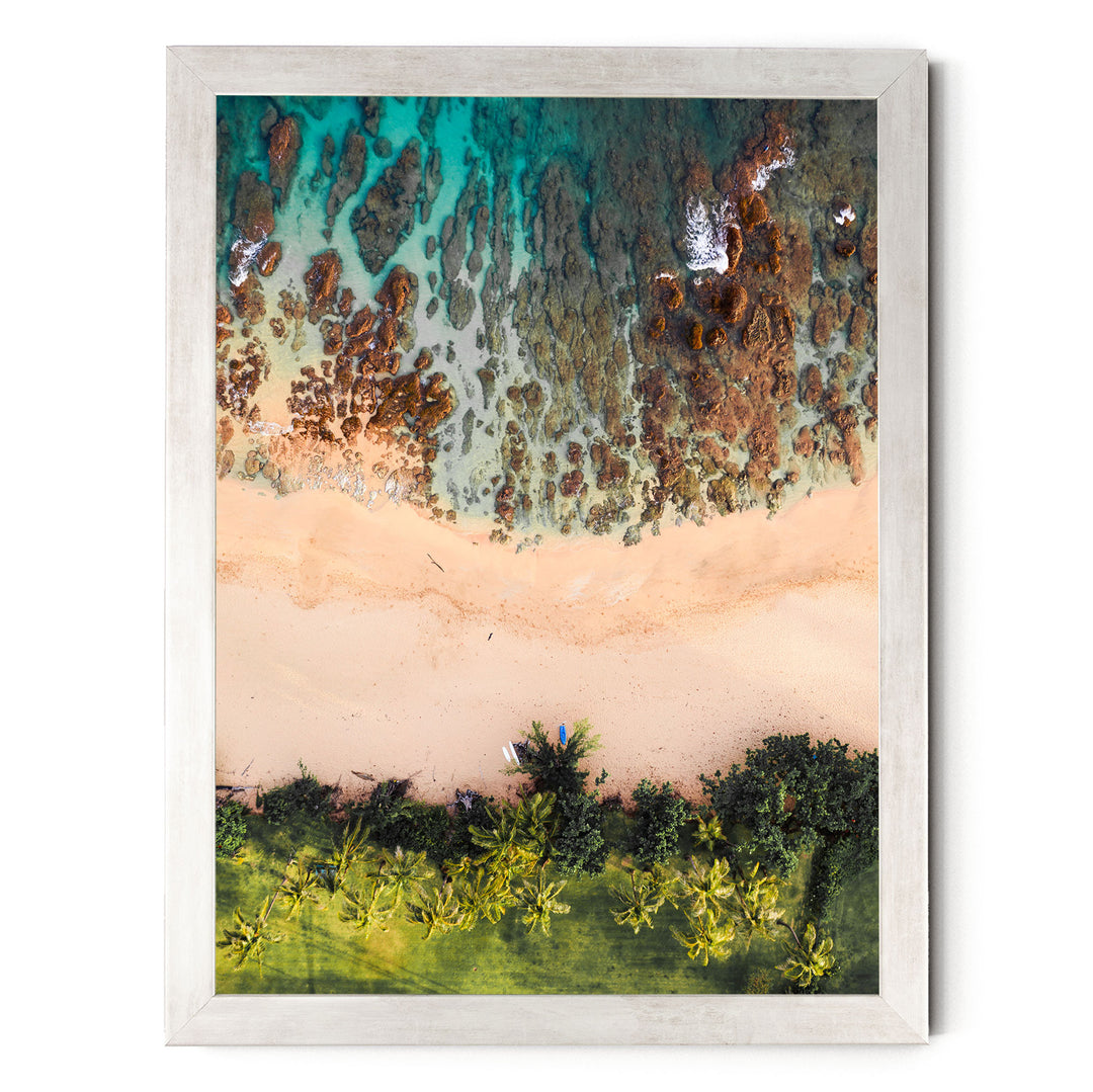 The image is an aerial view of a beach with a clear blue sky, green vegetation, and a sandy shoreline.