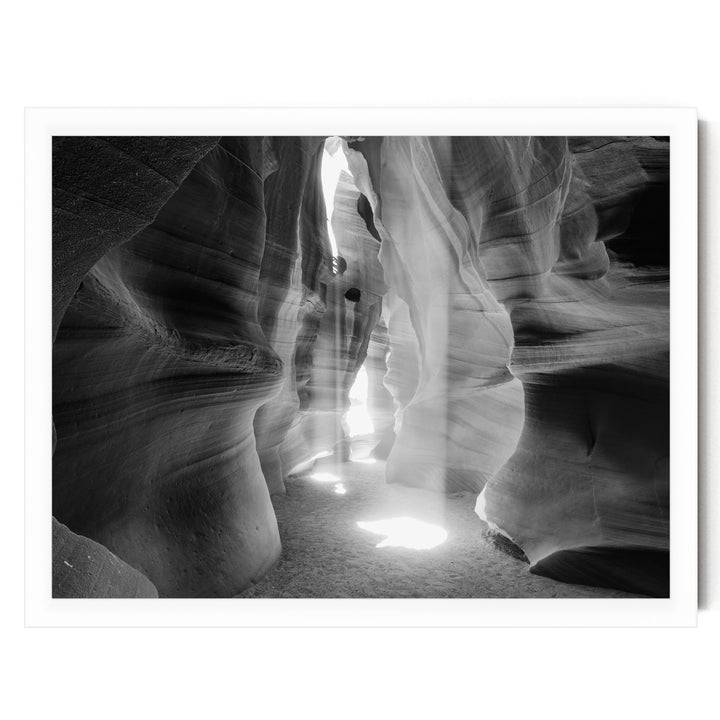A black and white image of a narrow passage in a rock formation, with sunlight streaming through the opening and casting shadows on the walls.