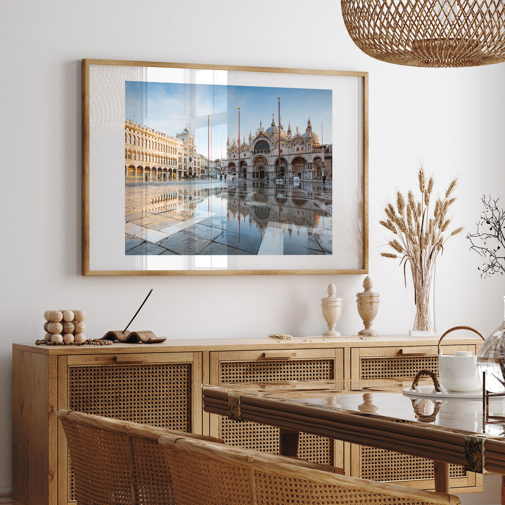 a large framed photograph of the Doge's Palace in Venice, Italy, hanging on a wall above a wooden cabinet. The photograph captures the palace's reflection in a large pool of water, creating a serene and picturesque scene. The photograph is framed in a gold frame, adding a touch of elegance to the display. The room has a rustic, natural aesthetic with woven furniture and a woven light fixture hanging from the ceiling.