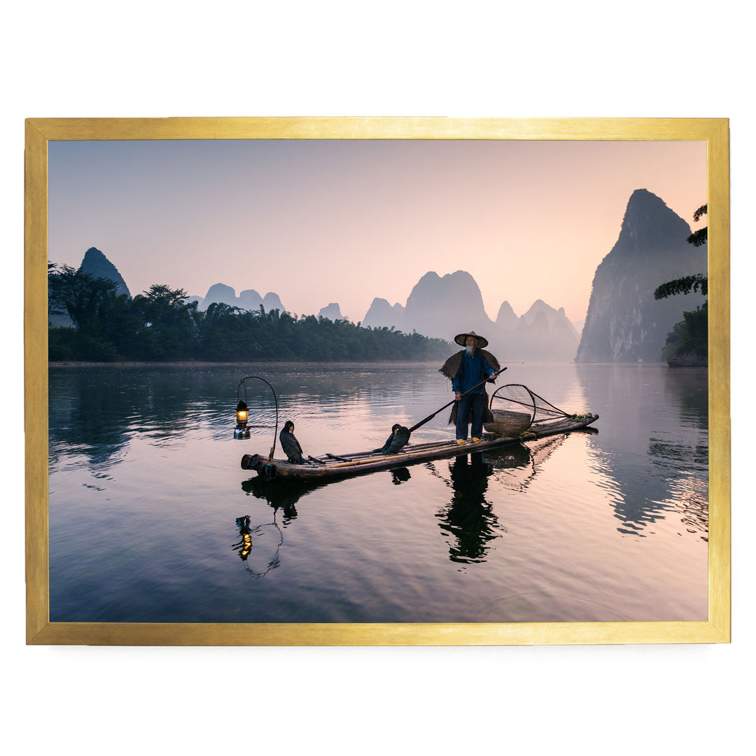 A person in a traditional Chinese outfit is standing on a long wooden boat, paddling across a calm river at sunset.