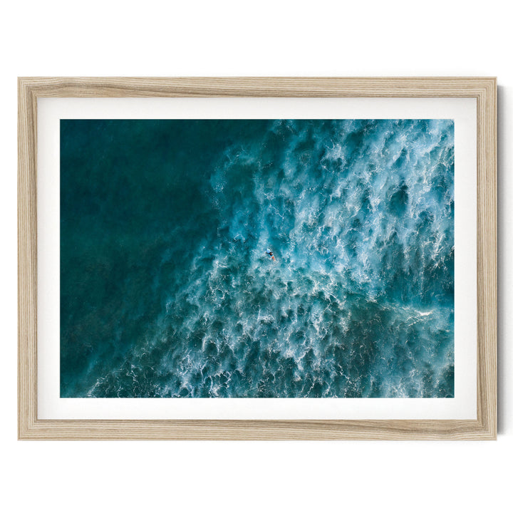 The image is a framed aerial view of a large body of water, likely an ocean, with waves and a person visible in the water.