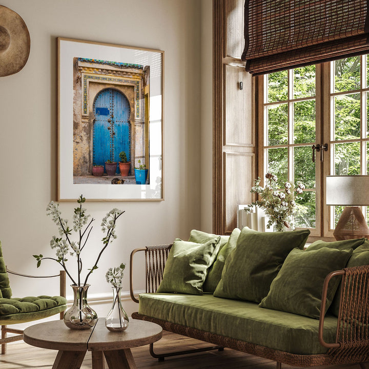 a cozy living room with a large window overlooking a lush green outdoor scene. The room is furnished with a green sofa, a coffee table, and a framed picture of a blue door with a green shutters. The room is decorated with potted plants and vases, creating a warm and inviting atmosphere.