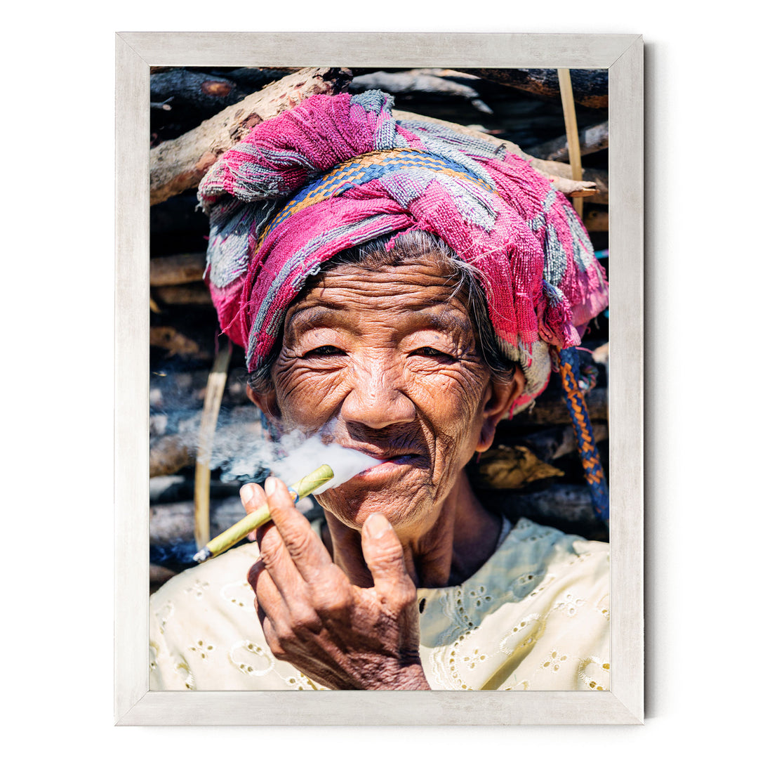 An elderly woman wearing a colorful headscarf and holding a lit cigarette, with a pile of wood in the background.