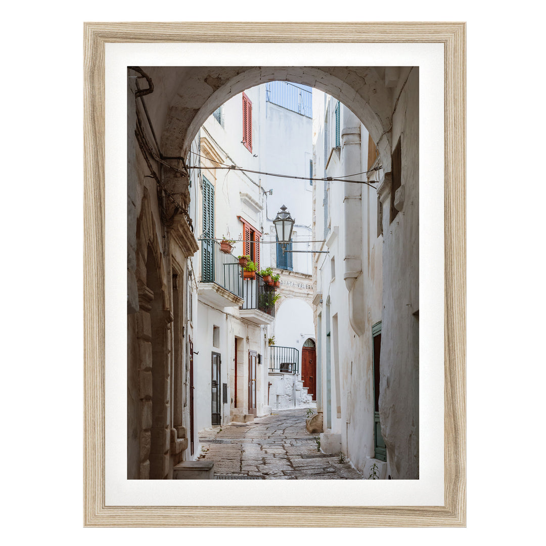 A narrow cobblestone street lined with white buildings and colorful shutters, leading to a balcony with potted plants.