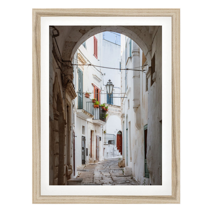 A narrow cobblestone street lined with white buildings and colorful shutters, leading to a balcony with potted plants.
