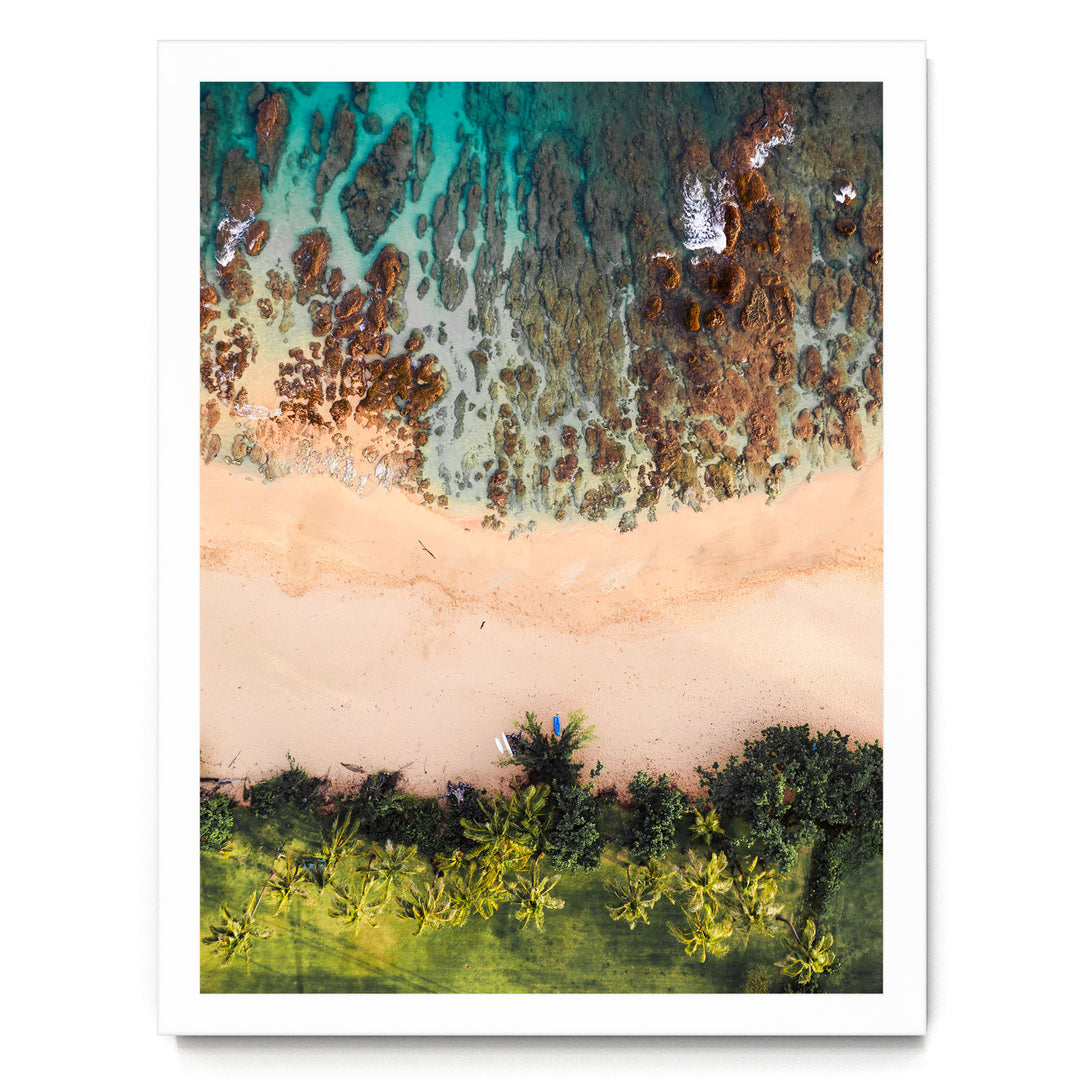 The image is an aerial view of a beach with a mix of rocky and sandy areas, surrounded by lush greenery and palm trees.
