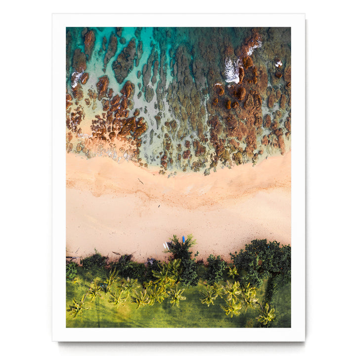 The image is an aerial view of a beach with a mix of rocky and sandy areas, surrounded by lush greenery and palm trees.