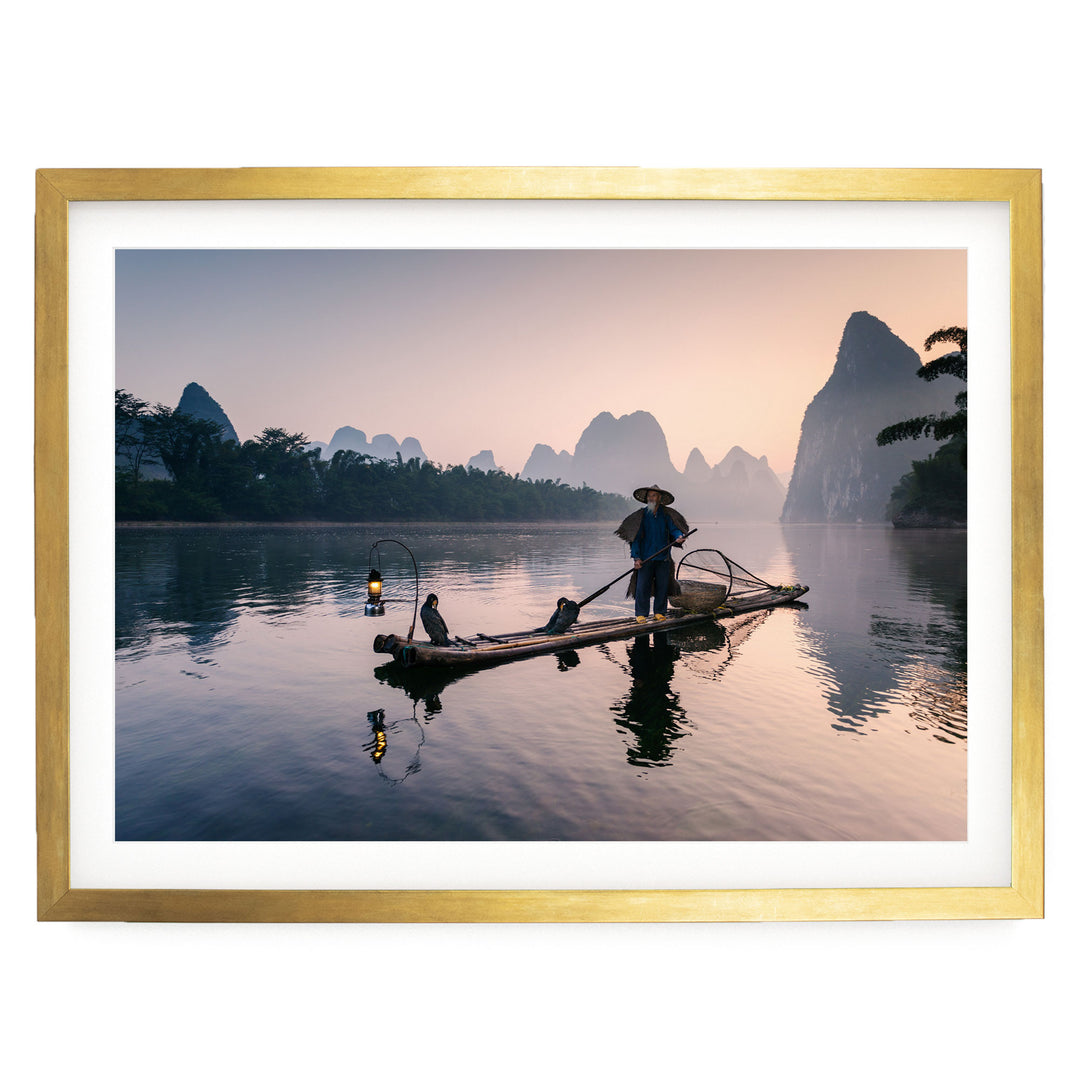 A person stands on a long wooden boat, paddling across a calm body of water surrounded by mountains and trees.