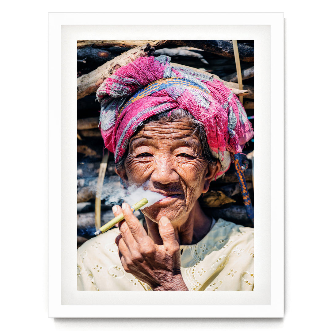 An elderly woman wearing a pink headscarf and a white blouse is shown smoking a cigarette, with a pile of wood in the background.