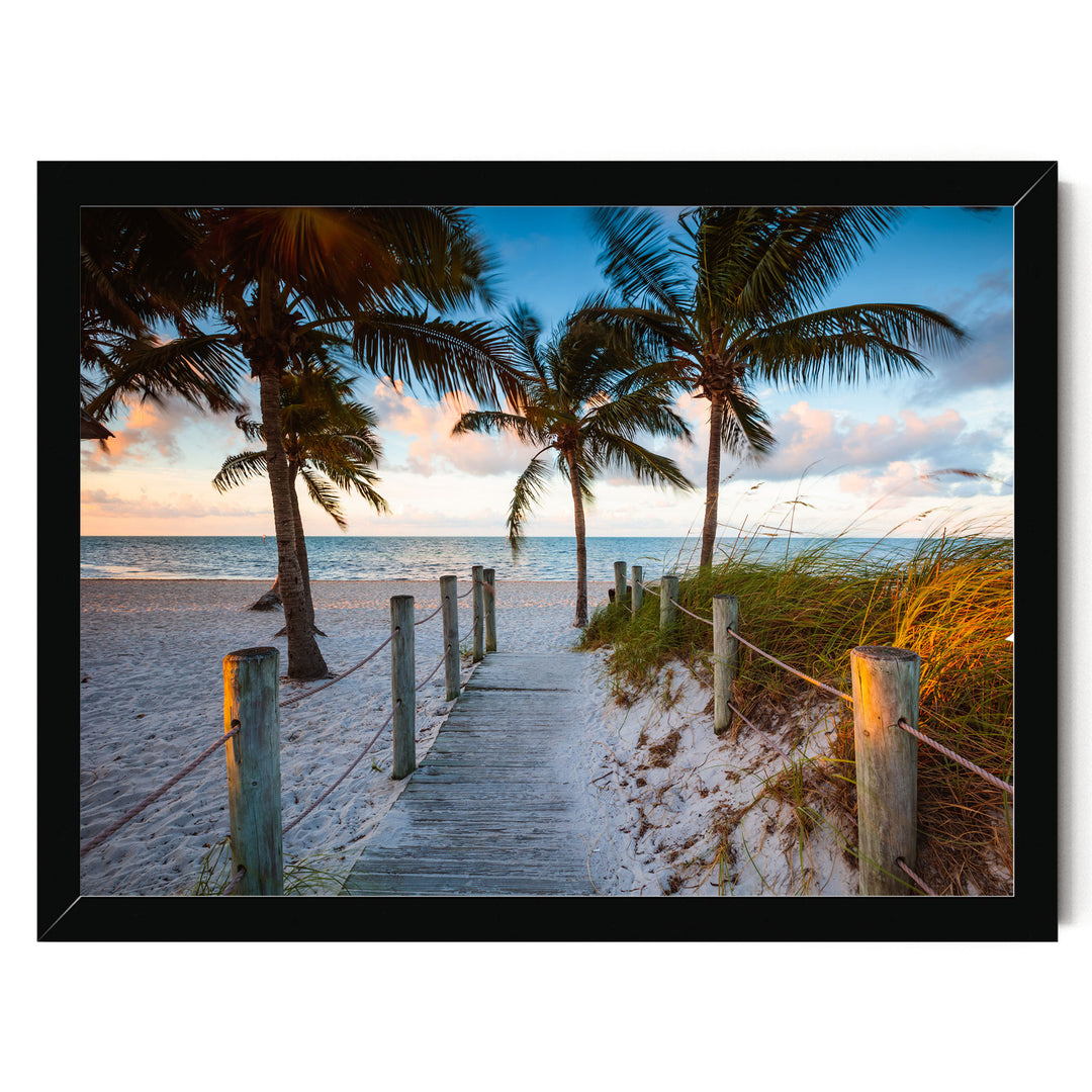 A wooden walkway leads to a sandy beach, with swaying palm trees and a picturesque sunset in the background.