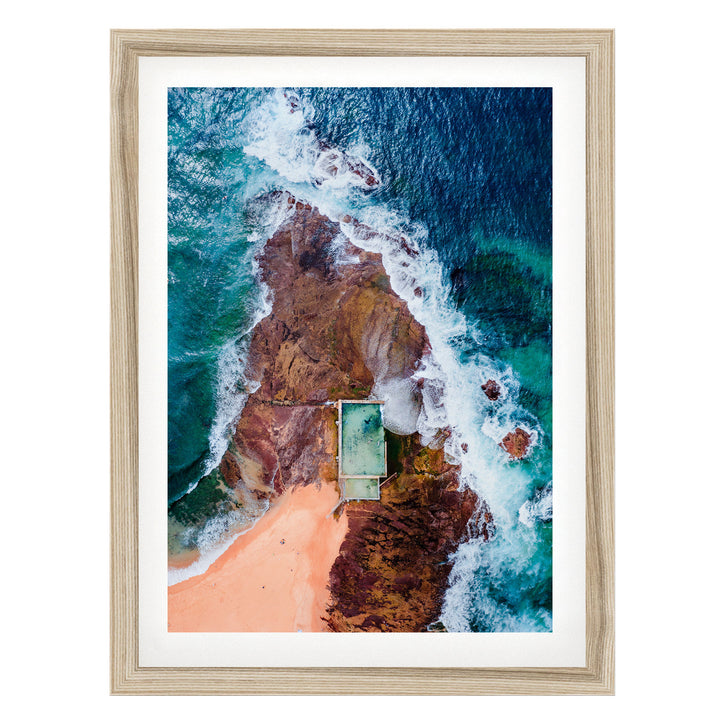 The image is an aerial view of a beach with a swimming pool, surrounded by rocky cliffs and crashing waves.