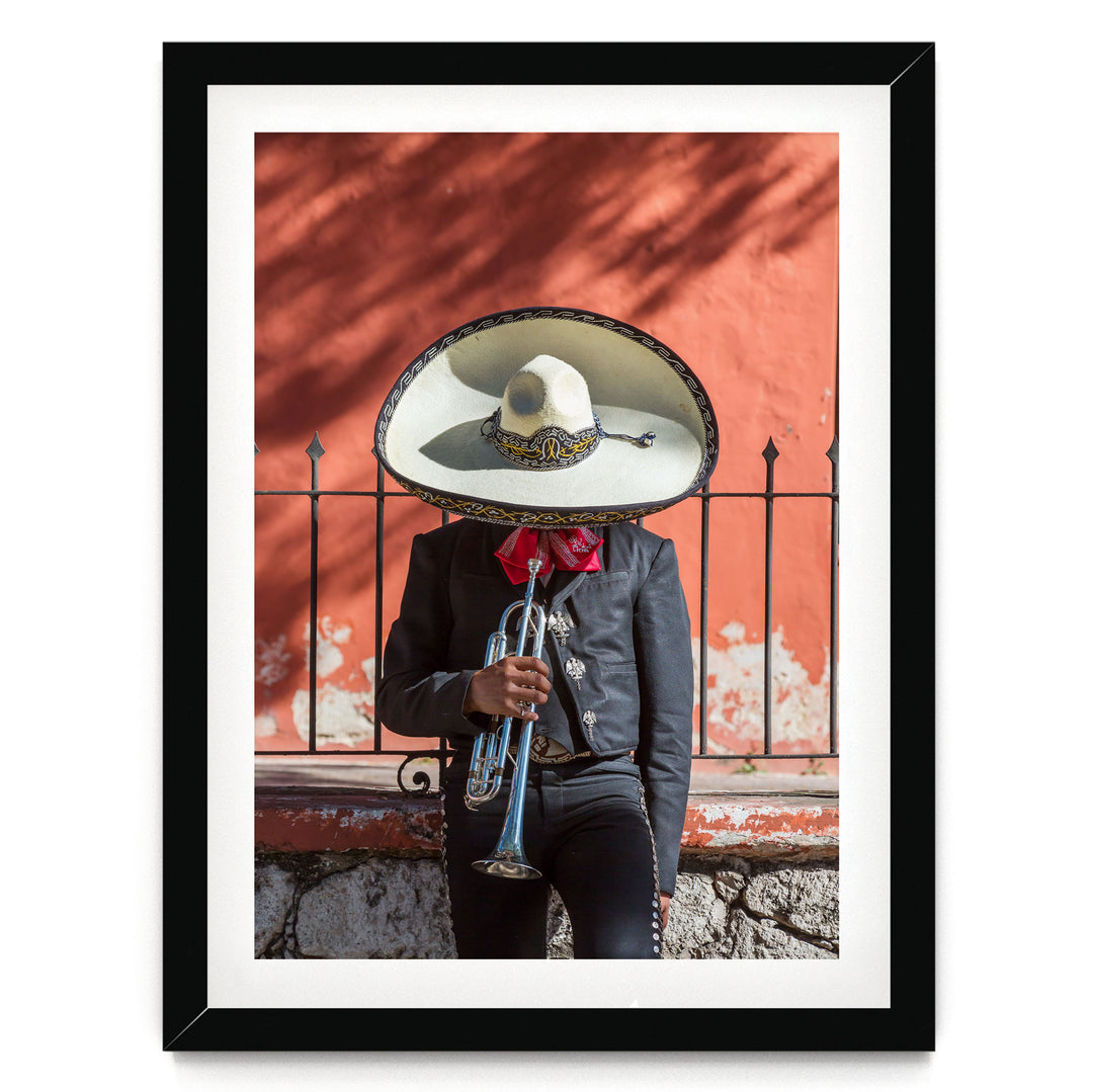A person wearing a sombrero and a black jacket is playing a trumpet in front of a red wall.