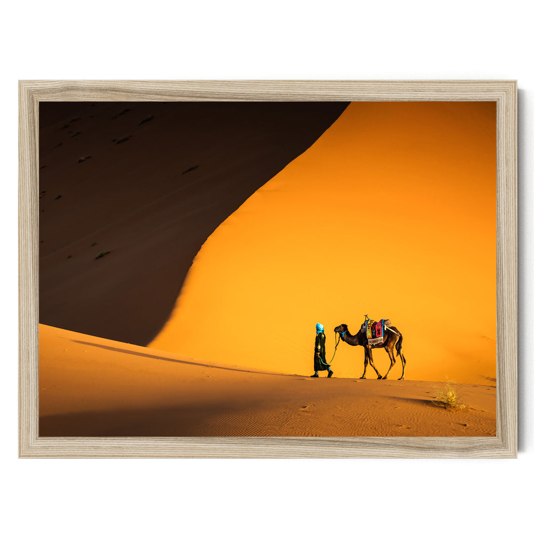 A person in a green outfit walks alongside a camel in a desert setting, with a large sand dune in the background.