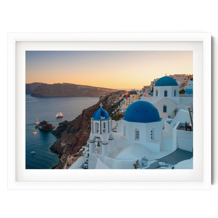 a picturesque coastal town with white buildings and blue domes, overlooking the ocean. The town is situated on a cliff, with a clear blue sky and a warm sunset in the background.