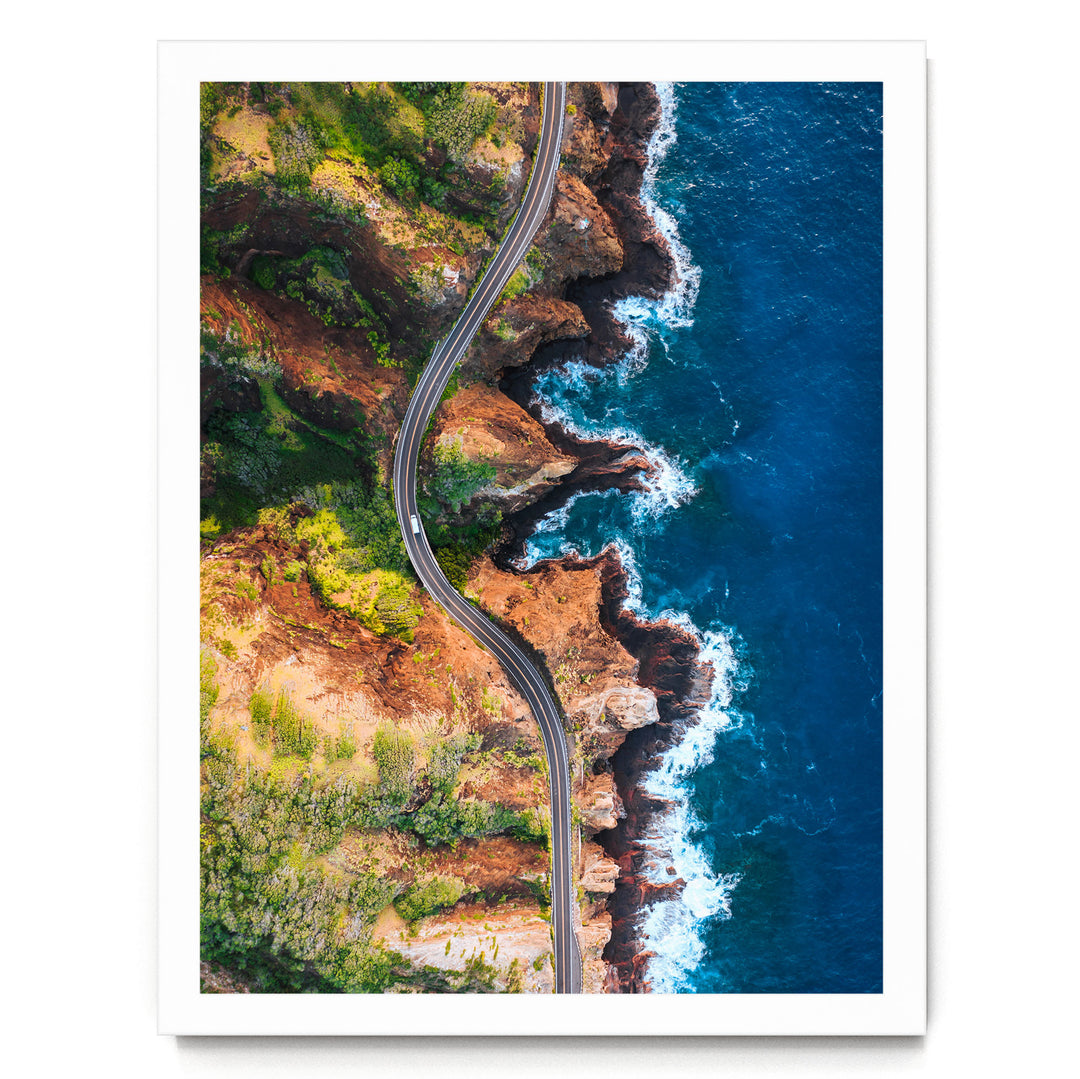 An aerial view of a winding road cutting through a rugged coastline with cliffs, lush vegetation, and the ocean in the background.