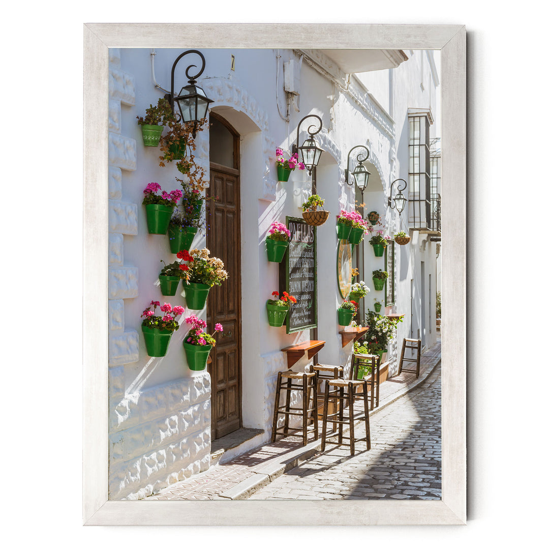 A charming street scene with white buildings adorned with hanging flower pots and green planters, creating a vibrant and inviting atmosphere.