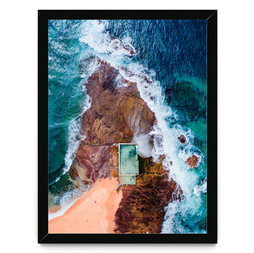 The image is an aerial view of a beach with a swimming pool, surrounded by rocky cliffs and crashing waves.