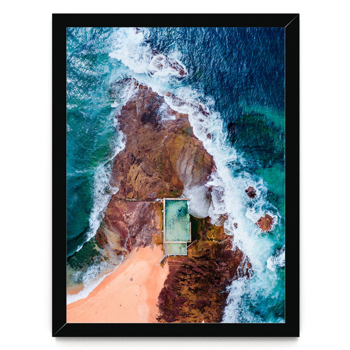 The image is an aerial view of a beach with a swimming pool, surrounded by rocky cliffs and crashing waves.