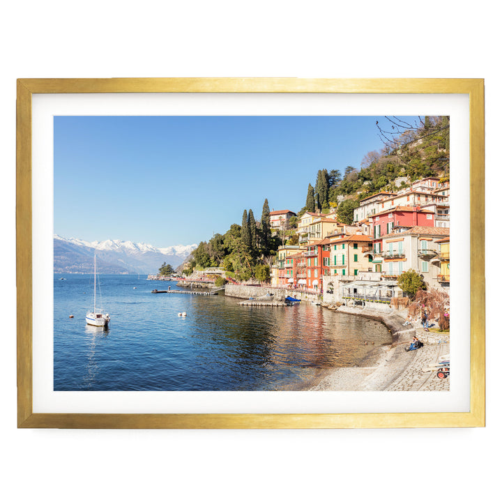 a picturesque coastal town with colorful buildings lining the shoreline, overlooking a serene lake. A sailboat is visible on the water, and the sky is a clear blue.