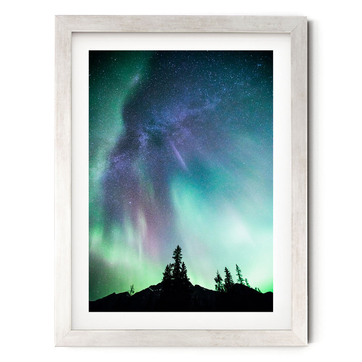 a stunning night sky with a vibrant display of the Aurora Borealis, or Northern Lights, illuminating the surrounding landscape. The silhouettes of trees can be seen in the foreground, adding a sense of depth and scale to the scene.