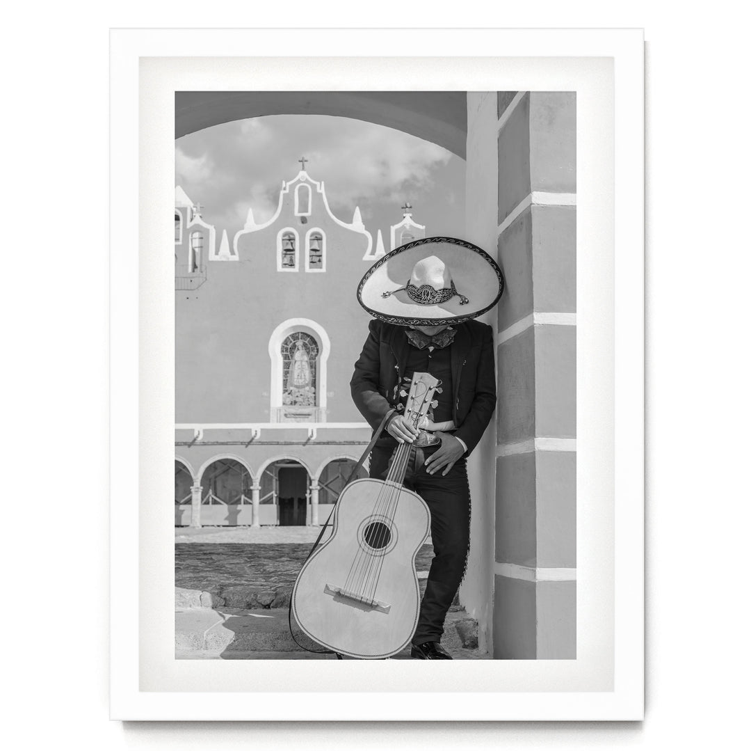 A person in a sombrero and traditional Mexican attire is playing a guitar in front of a building with arched windows and a cross on top.