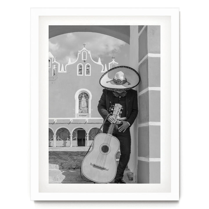 A person in a sombrero and traditional Mexican attire is playing a guitar in front of a building with arched windows and a cross on top.