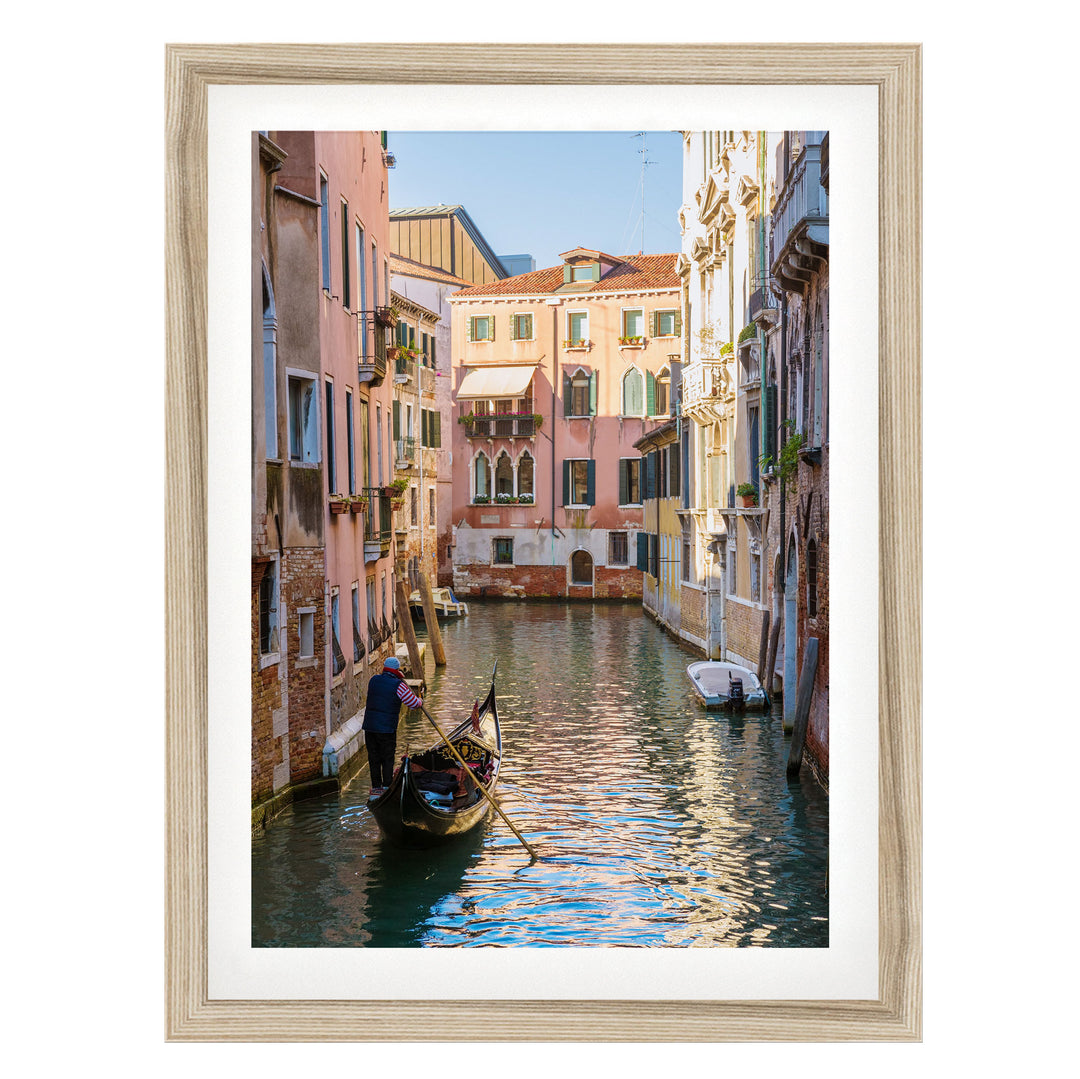 A gondolier navigates a traditional Venetian gondola through a picturesque canal, surrounded by colorful buildings and reflecting the clear blue sky.
