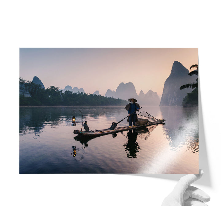 A person stands on a long wooden boat, paddling across a calm body of water surrounded by mountains and trees.