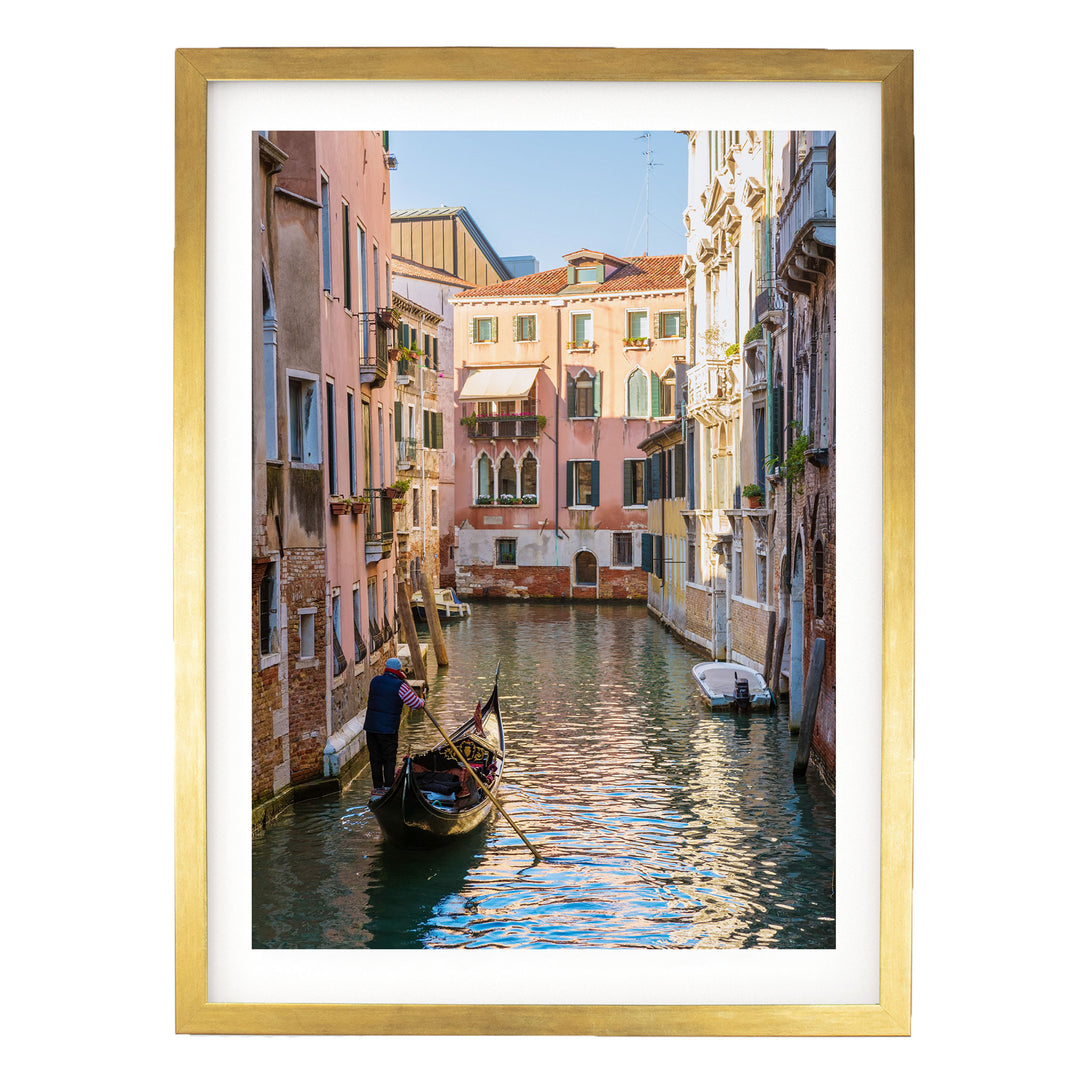 A gondolier in a traditional Venetian gondola navigates a picturesque canal in Venice, surrounded by colorful buildings and reflecting the clear blue sky.