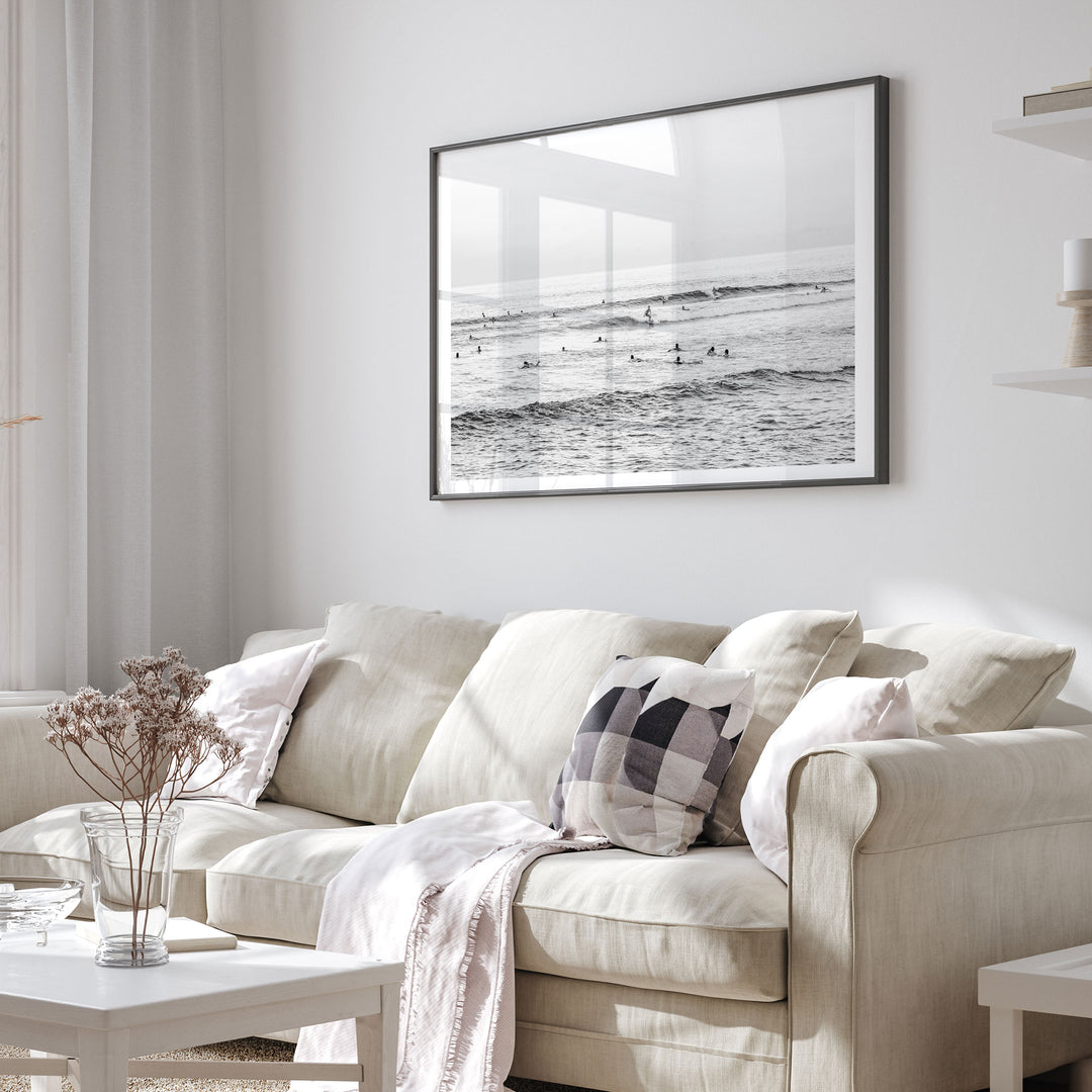 a cozy living room with a beige sofa adorned with a checkered throw pillow and a white blanket draped over it. Above the sofa, a framed black and white photograph of people surfing on a beach is displayed on the wall.