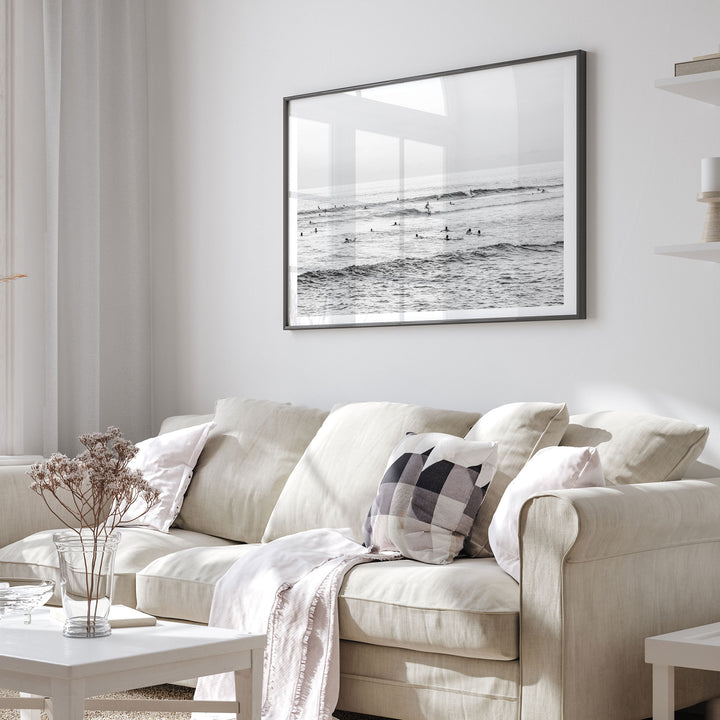 a cozy living room with a beige sofa adorned with a checkered throw pillow and a white blanket draped over it. Above the sofa, a framed black and white photograph of people surfing on a beach is displayed on the wall.