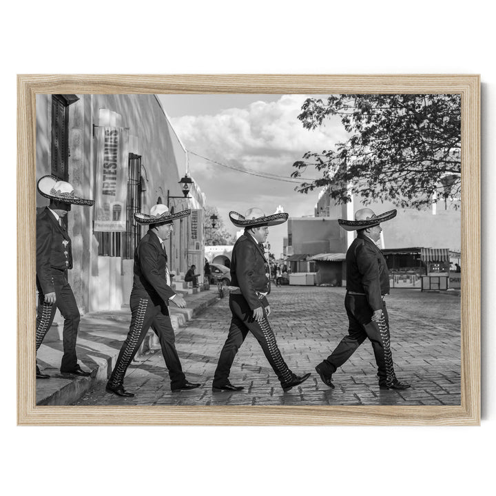 A group of men wearing sombreros and dark clothing are walking down a street, with a building and trees visible in the background.