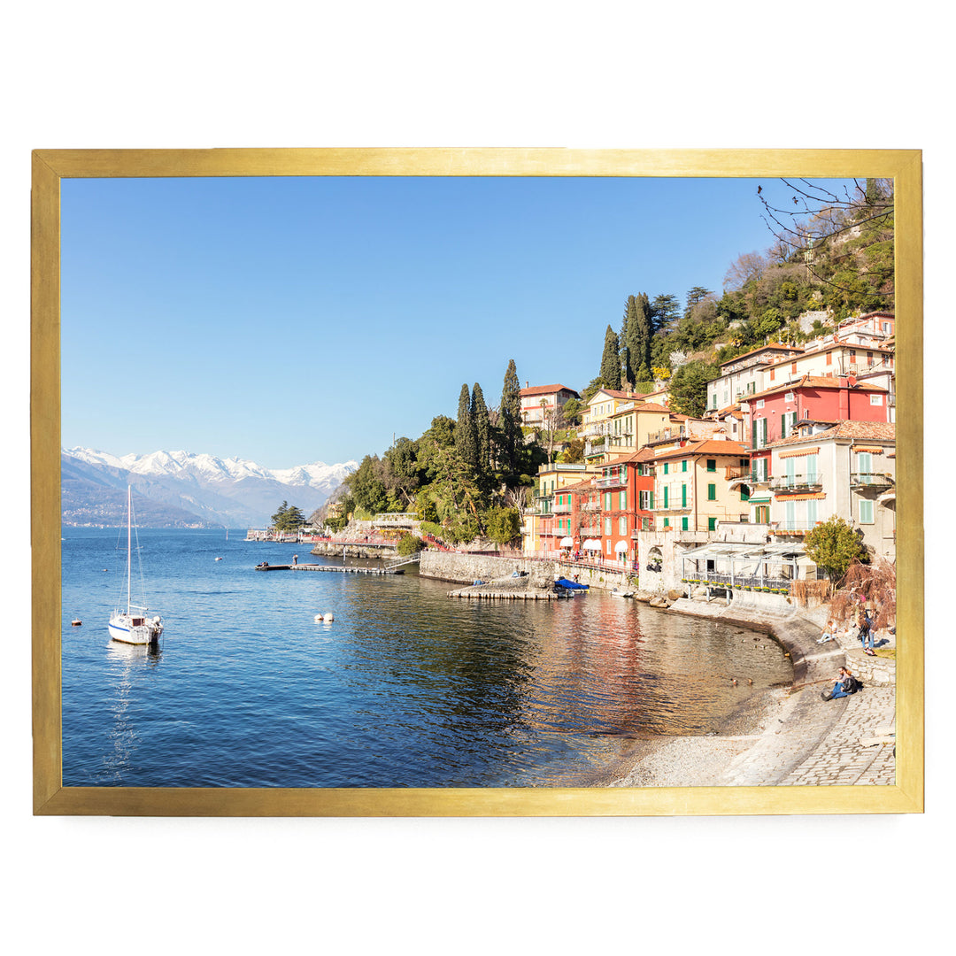 A picturesque coastal town with colorful buildings nestled along the shoreline, overlooking a serene lake. A sailboat is visible on the water, and the surrounding landscape is dotted with trees and mountains in the distance.