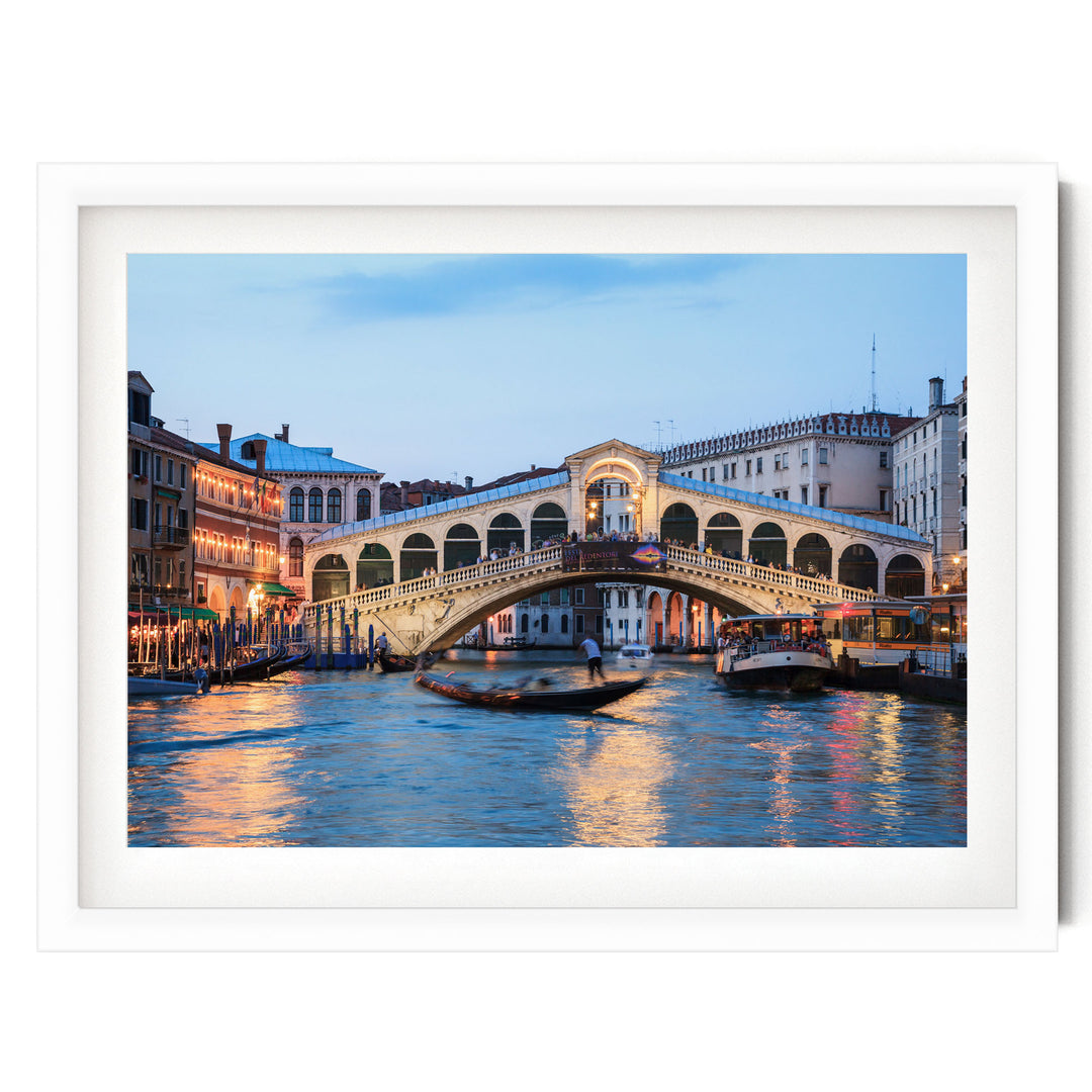 a picturesque scene of the Rialto Bridge in Venice, Italy, with its distinctive white stone arch spanning the Grand Canal. The bridge is illuminated at dusk, creating a warm glow that reflects off the water. The canal is filled with boats, adding to the lively atmosphere of the scene. The image is framed within a white border, highlighting the beauty of the landmark and the surrounding architecture.