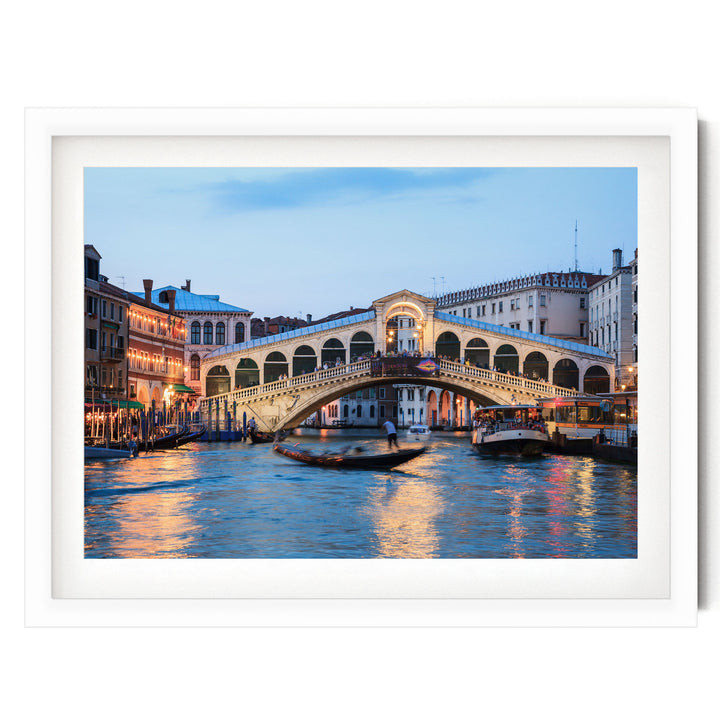 a picturesque scene of the Rialto Bridge in Venice, Italy, with its distinctive white stone arch spanning the Grand Canal. The bridge is illuminated at dusk, creating a warm glow that reflects off the water. The canal is filled with boats, adding to the lively atmosphere of the scene. The image is framed within a white border, highlighting the beauty of the landmark and the surrounding architecture.