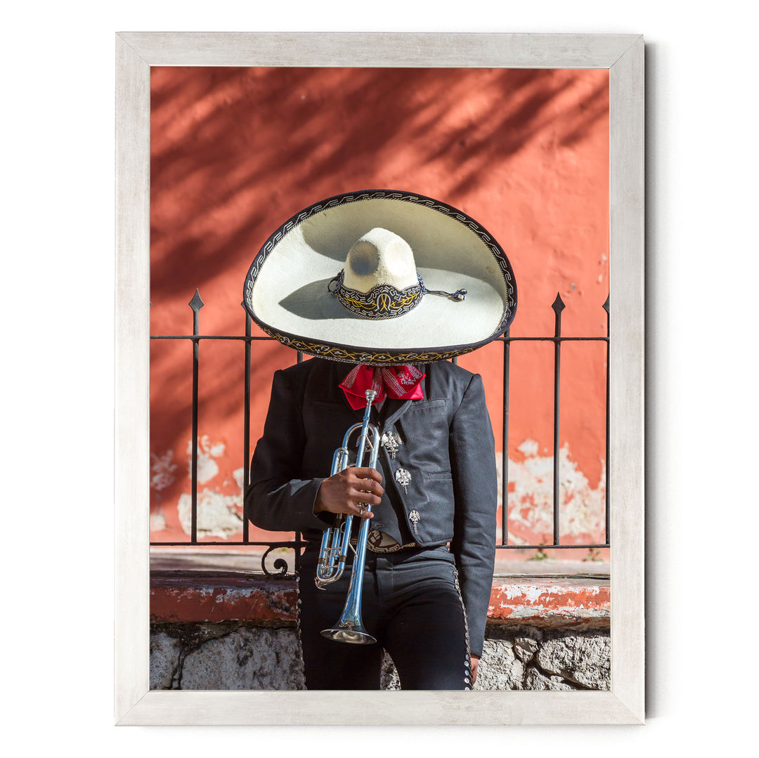 A person wearing a sombrero and a black jacket is playing a trumpet in front of a red wall.