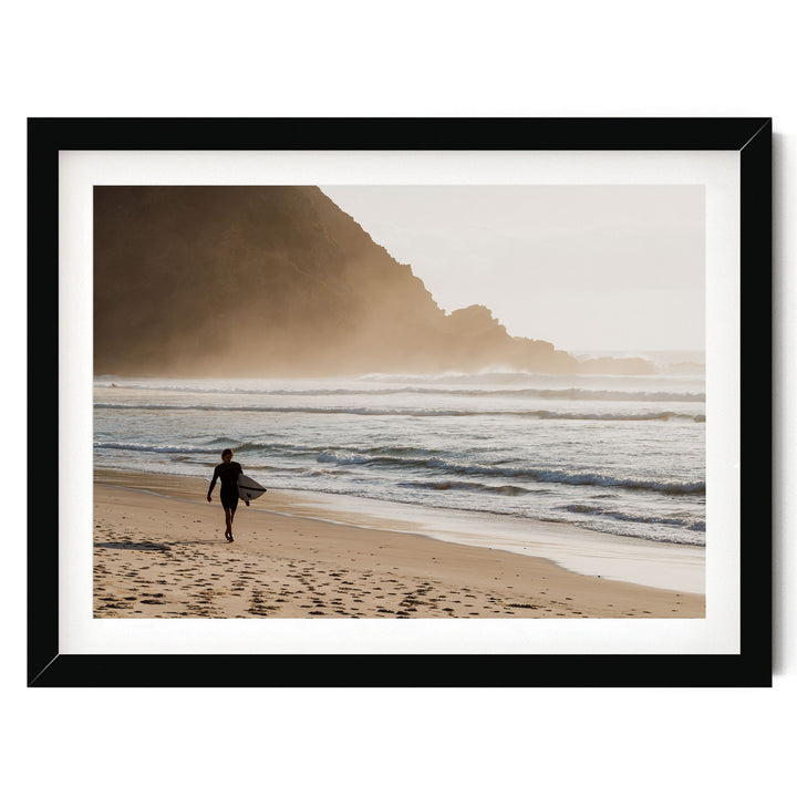 A person in a wetsuit carrying a surfboard walks along a sandy beach, with a rocky cliff and ocean in the background.