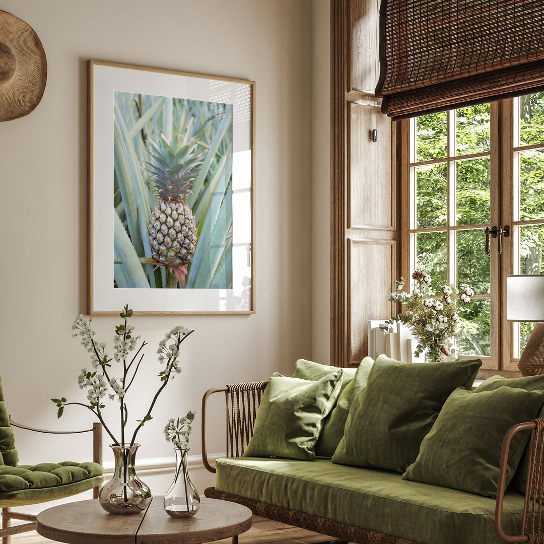 a cozy living room with a large window overlooking lush greenery outside. The room is adorned with a green sofa, a wooden coffee table, and a framed picture of a pineapple. The room is further decorated with potted plants and vases, creating a serene and inviting atmosphere.