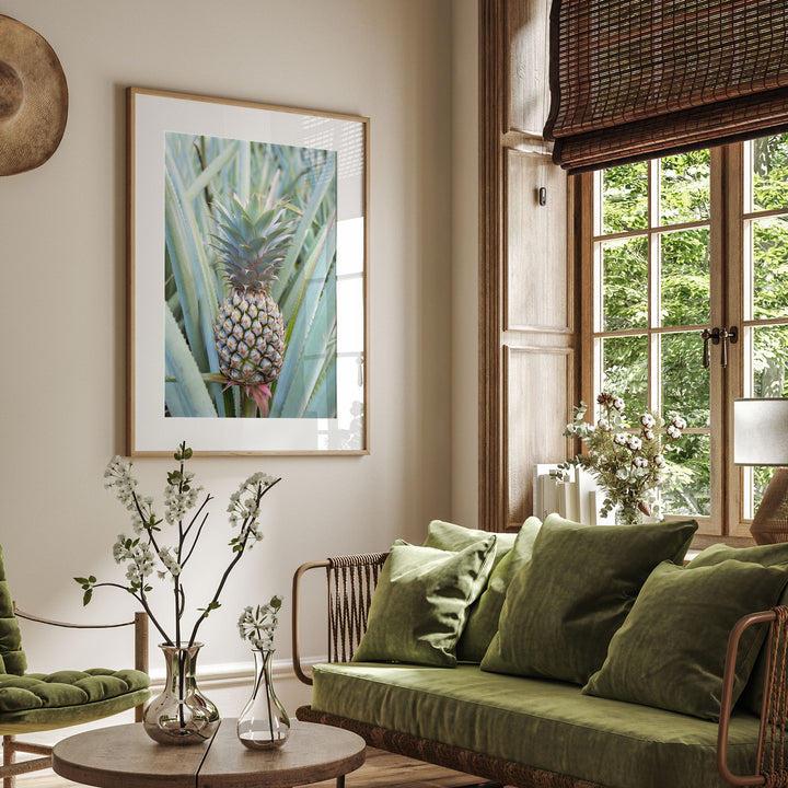 a cozy living room with a large window overlooking lush greenery outside. The room is adorned with a green sofa, a wooden coffee table, and a framed picture of a pineapple. The room is further decorated with potted plants and vases, creating a serene and inviting atmosphere.