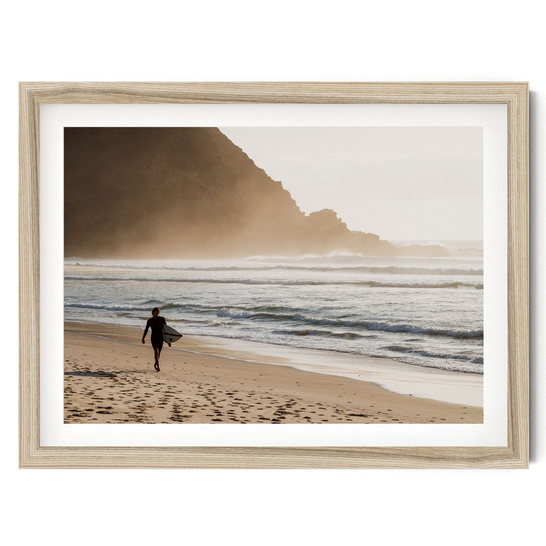 A surfer walks along the beach, carrying a surfboard, with a rocky cliff and ocean in the background.