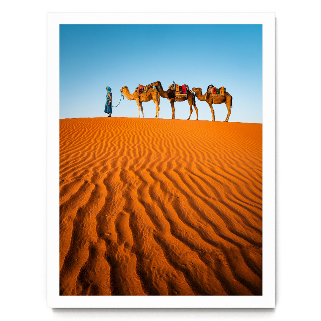 A person in a blue robe leads three camels across a vast, orange sand dune in a desert landscape.