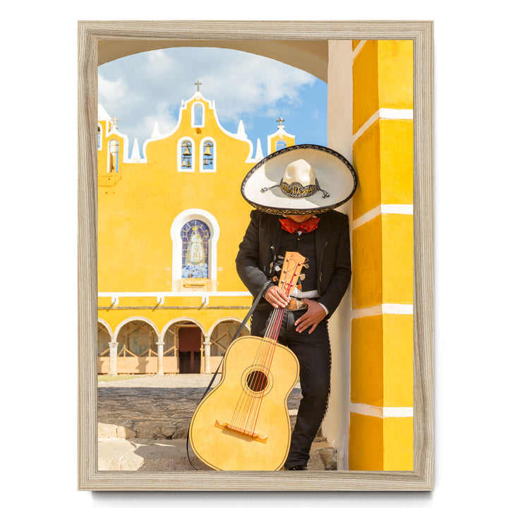 A person in a sombrero and traditional Mexican attire is playing a guitar in front of a yellow building with arched windows and a cross on top.