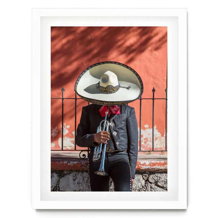 A person wearing a sombrero and a black jacket is playing a trumpet in front of a red wall.